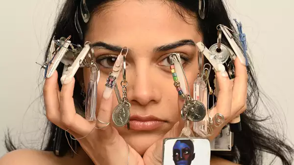 A person with decorated nails holding their face, featuring various keychains and trinkets attached to their nails.