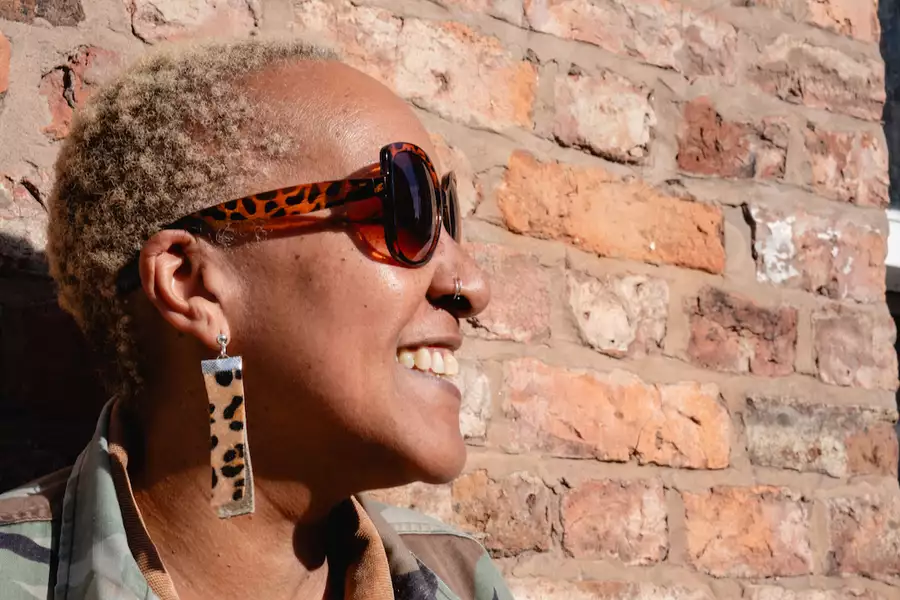 Andrea Trout smiling and looking to the side, wearing leopard print sunglasses and earrings. They are stood in front of a brick wall.
