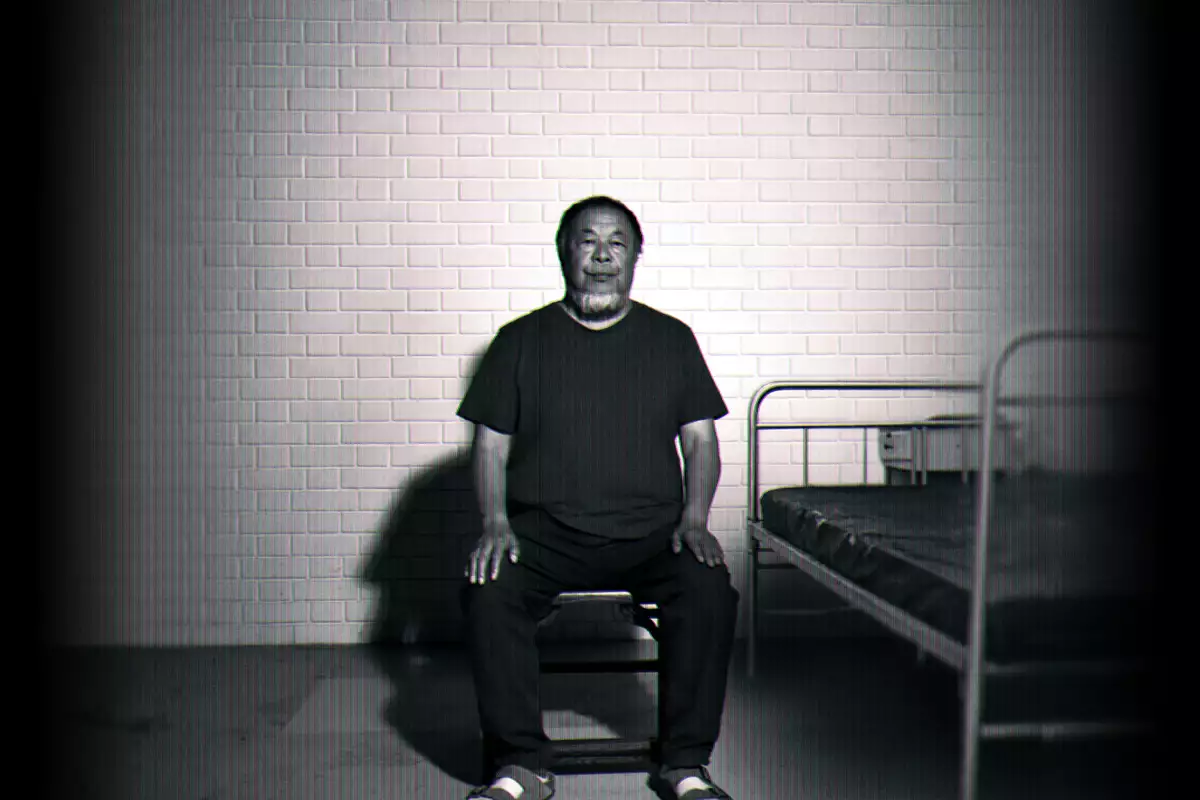 Black-and-white image of Ai Weiwei sitting on a chair beside a simple metal bed against a brick wall.