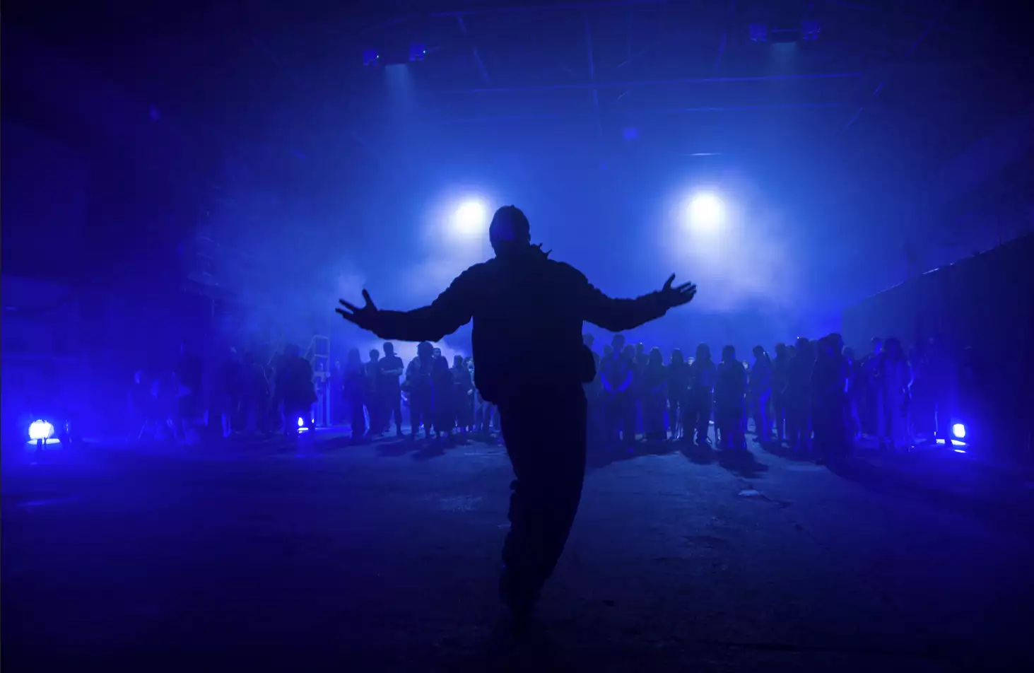 A photo of warehouse industrial space lit in blue light with a person standing in front of a crowd with his arms wide