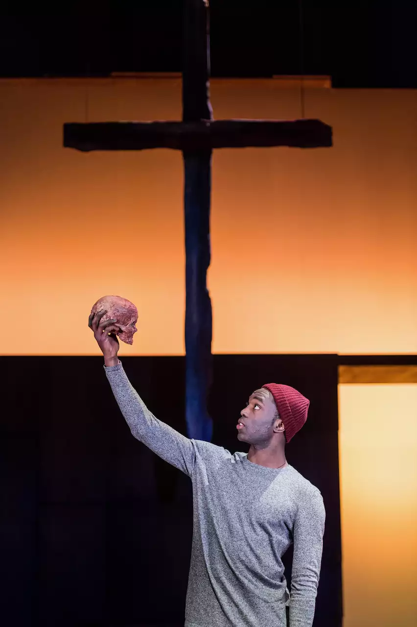 Paapa Essiedu as Hamlet wearing a grey top and red beanie hat. He is holding a skull in the air with one hand and looking at it. Behind him is a cross on a burnt-orange background.