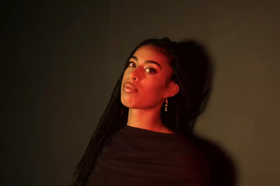 Victoria Jane in a black dress with braided hair, lit by red-toned lighting against a dark background.