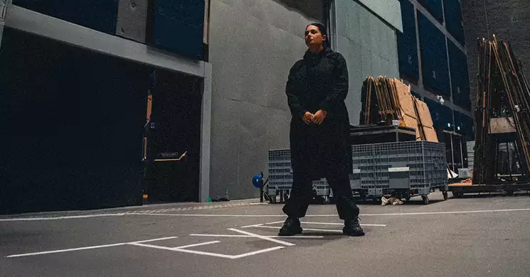 Hannah Laing dressed in black, standing in the Warehouse surrounded by equipment and stacked materials.