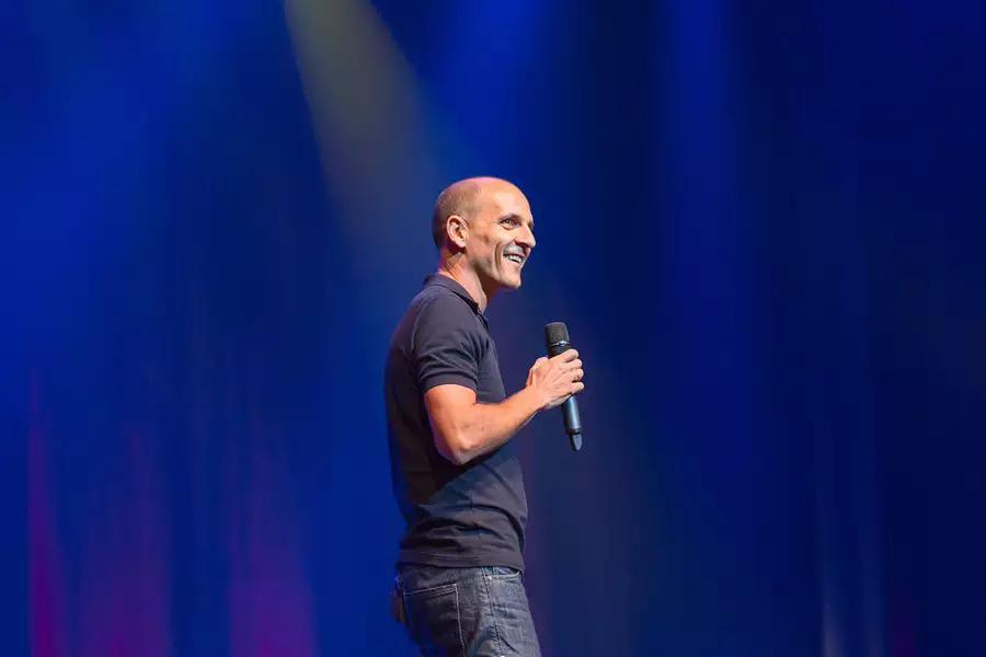 Carl Barron smiling and holding a microphone on stage, with blue curtains behind him