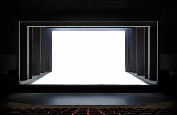 A theater stage with a dramatic, modern design is shown, featuring black framing and a stark white, illuminated backdrop. The contrast between the dark seating area and the bright stage creates a striking visual effect.