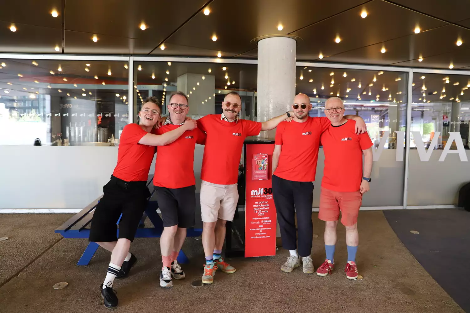 Five people in red t-shirts smiling outside Aviva Studios with the piano
