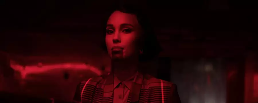 Kaneza Schaal in a dimly lit room bathed in intense red light, wearing a patterned jacket. The dramatic lighting and shadows create a cinematic atmosphere.