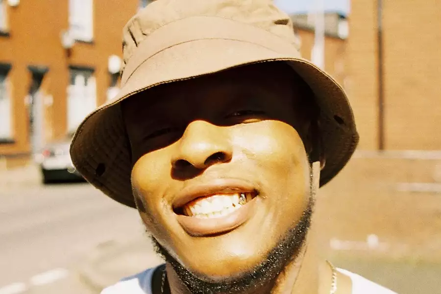 Close-up photo of Grumpy smiling wearing a light brown bucket hat. The background shows blurred terraced houses.
