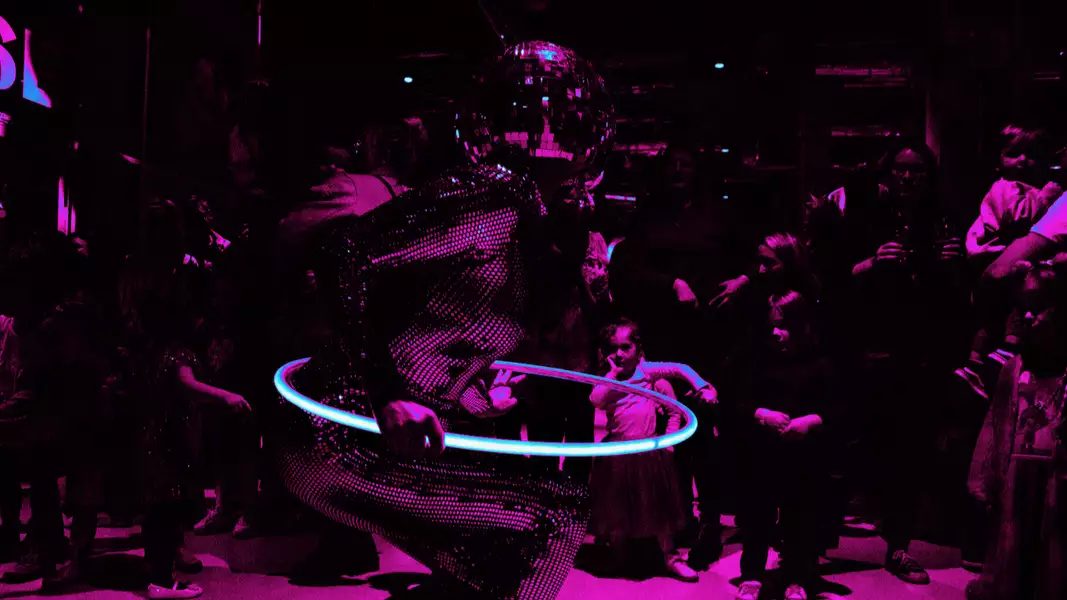 A performer in a disco-ball helmet and sparkling outfit dances with a glowing hula hoop under vibrant magenta lighting, surrounded by an audience.