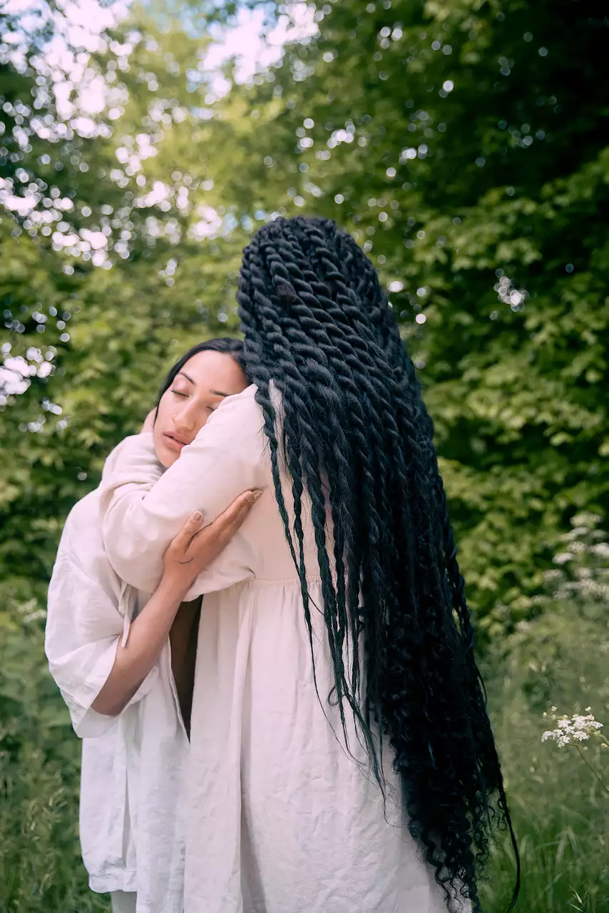 Two people embracing – both wearing flowing white clothes. One is turned away from the camera with long black hair. The other has their eyes closed. The background includes grass and trees out of focus.