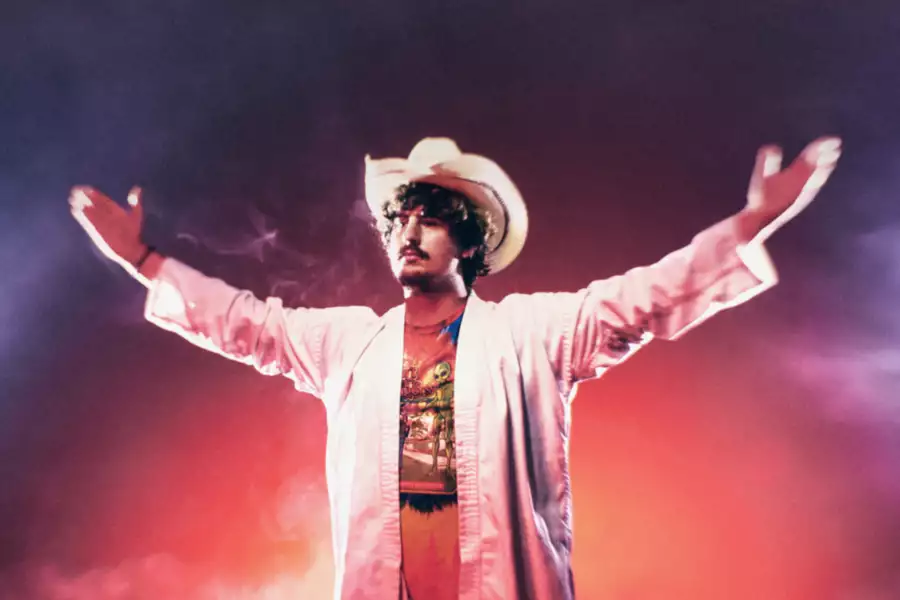 A man in a white suit and cowboy hat stands with arms outstretched, surrounded by dramatic red and blue lighting and thick mist.