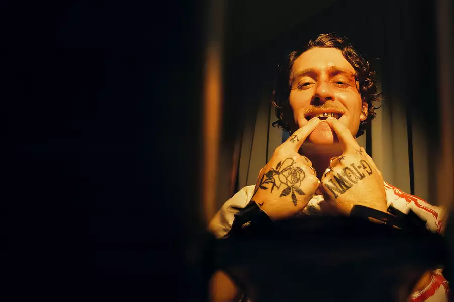 Photo of Kettama on a dark brown, shadowed background. His hands are tattooed and he is pointing at his metal teeth.