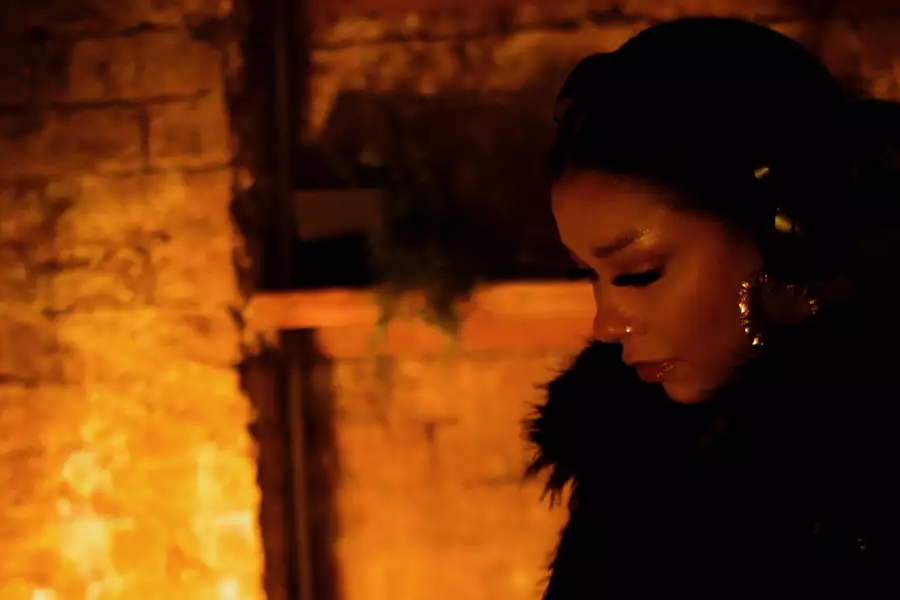 Katbrownsugar looking down, wearing a black top and gold earrings. The lighting is dim.