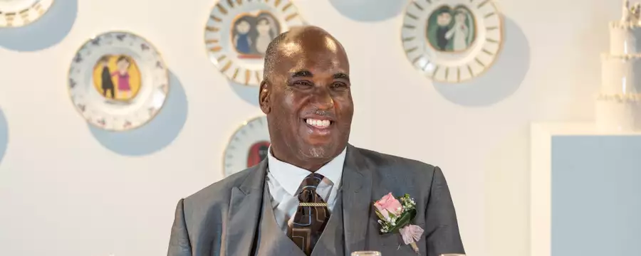 Horace Lindezey in a suit with a pink rose sits at a table with champagne glasses, in front of a wall decorated with illustrated plates.