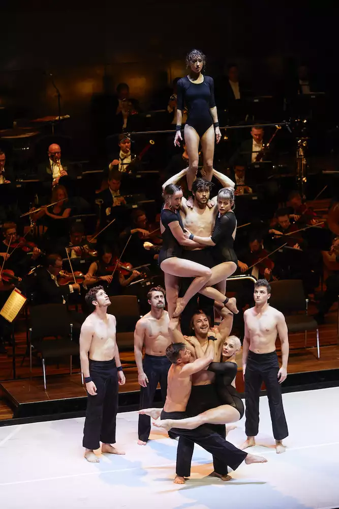 Circa acrobats stood on top of one another on stage, performing in front of a live orchestra