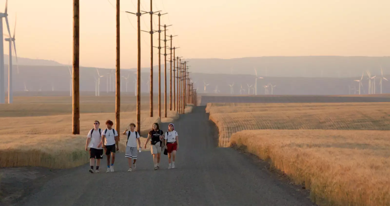 Still from Gasoline Rainbow. Five young people walking on an empty road with miles of empty fields, hills and wind turbines behind them