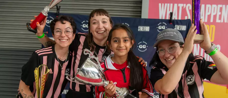 Three members of Girl Gang wearing football kits and posing with a child who is holding a trophy