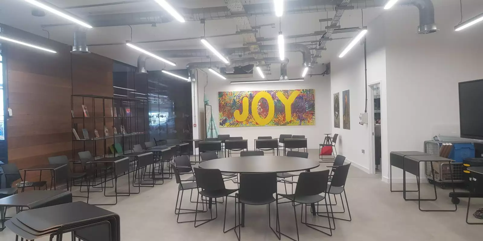 Photo of the Lab at Aviva Studios, showing an empty room with chairs, round tables and a colourful painting featuring the word 'JOY' on the wall