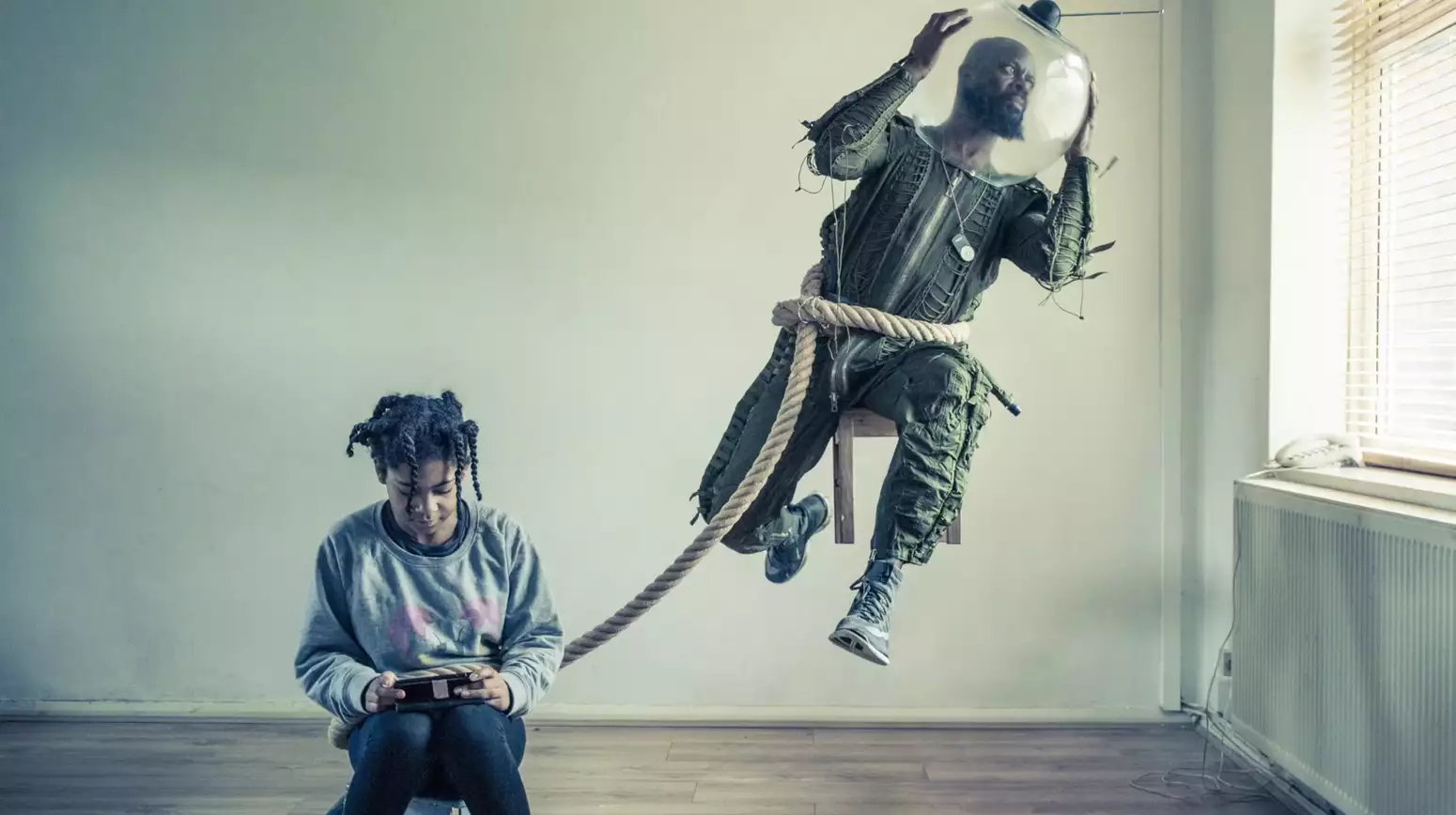 A photograph by Benji Reid, showing Benji sat on a suspended chair dressed in a space suit, while a young girl holds on to a rope round his waist