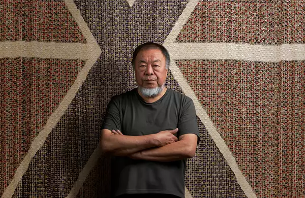 Ai Weiwei stood with arms folded wearing a black t-shirt, in front of a giant flag made of buttons