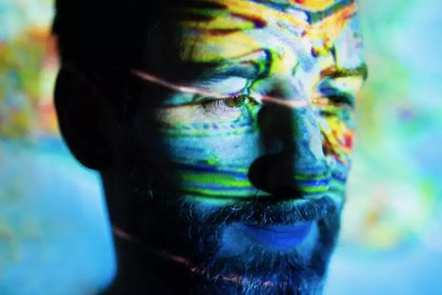 Max Cooper's face is illuminated with vibrant, multicoloured patterns projected onto his skin, creating a surreal and abstract effect. The background is softly blurred with similar hues, enhancing the dreamlike atmosphere.