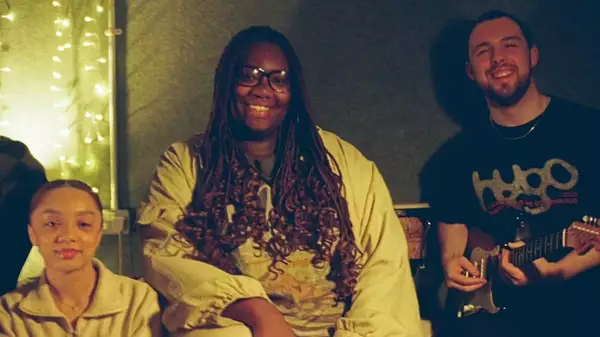 Three members of The Divines smiling. One holds a guitar. Behind them are softly lit fairy lights.