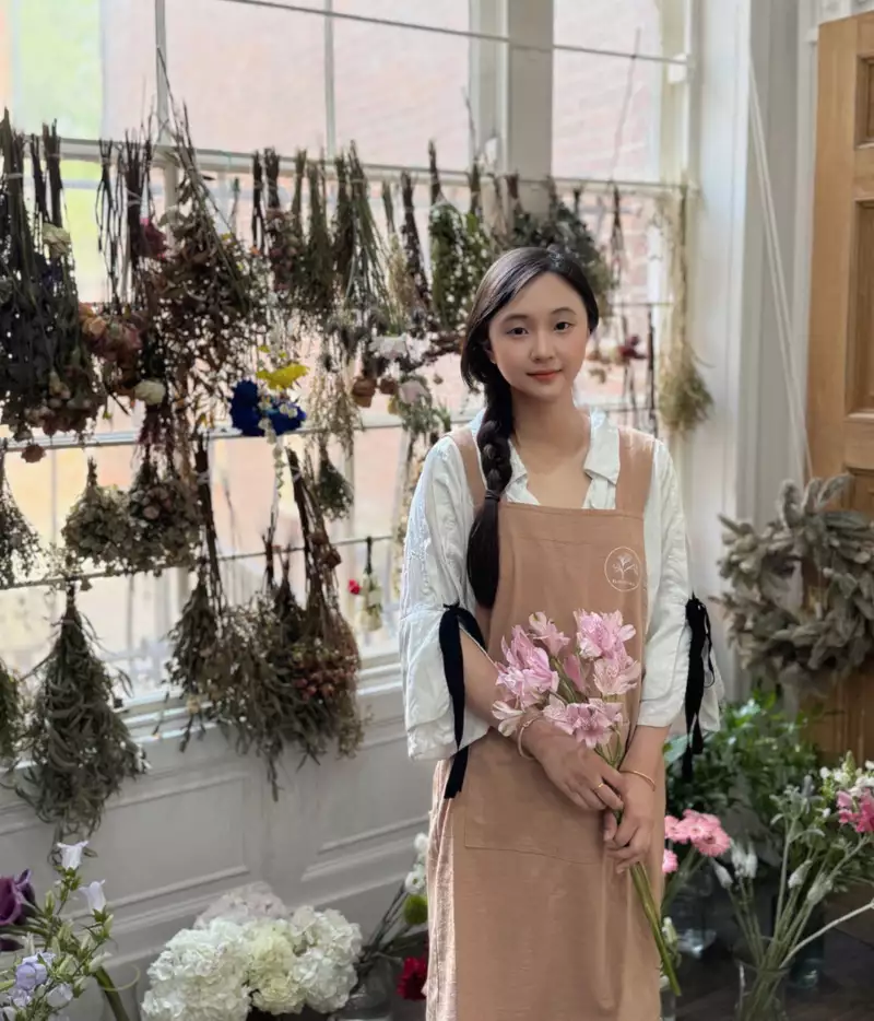 Kiki smiling in her art studio, wearing a light brown apron, holding flowers. There are many bunches of flowers behind her.