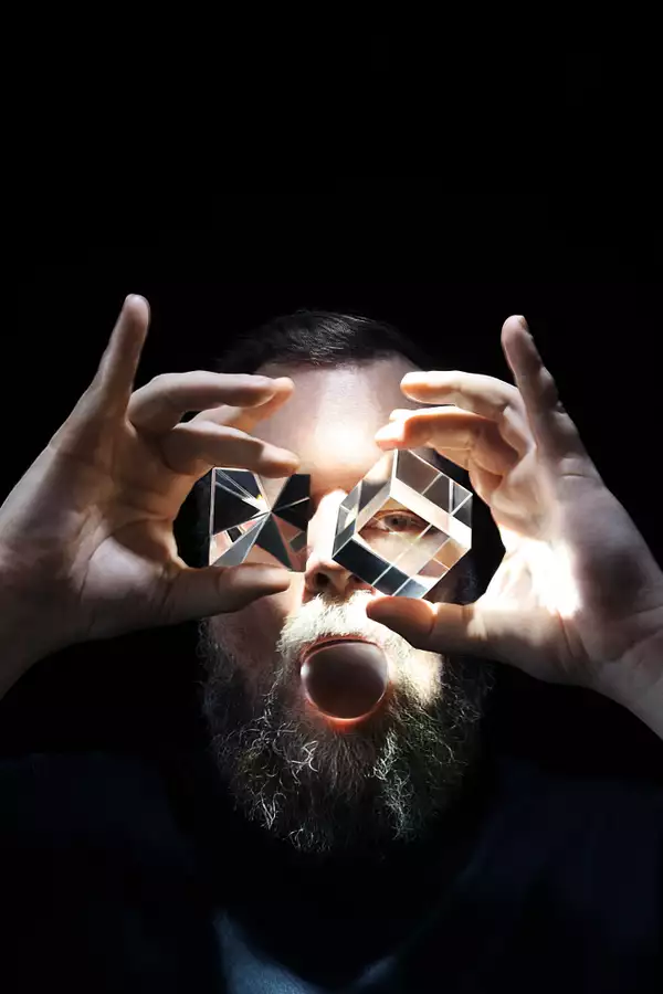 Headshot of singer-songwriter and musician John Grant holding a light in his mouth and crystal cubes over his eyes