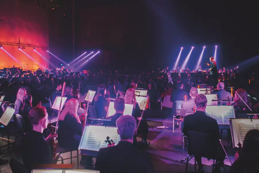 The BBC Philharmonic orchestra performing in the Warehouse. The lighting is pink and purple.  The audience are stood in the middle of the orchestra.