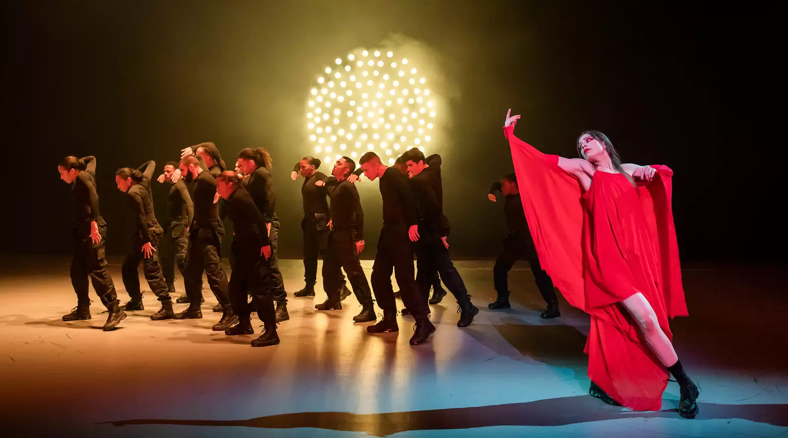 Dancer performing in a red dress with several dancers in black clothing, in front of a bright light in Free Your Mind