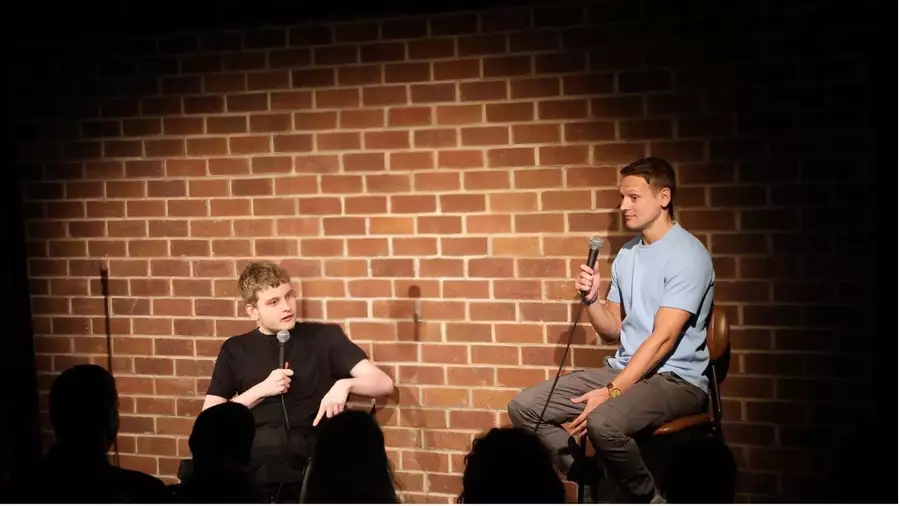 Wheels and the Legman performing on stage holding microphones. Behind them is a brick wall.