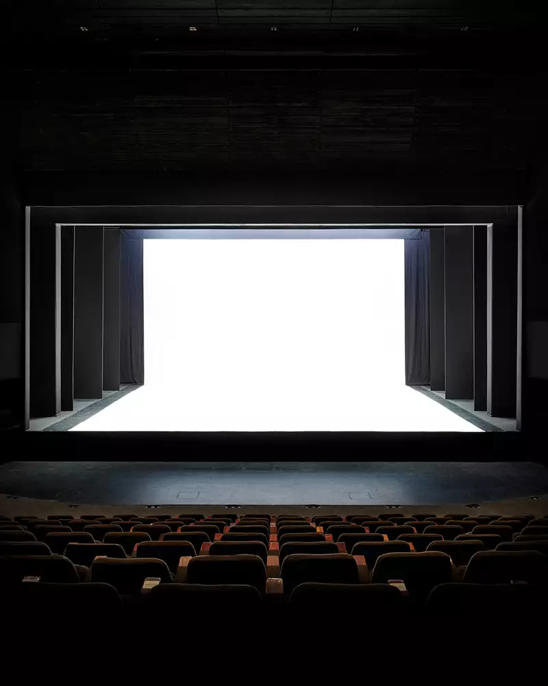 A theater stage with a dramatic, modern design is shown, featuring black framing and a stark white, illuminated backdrop. The contrast between the dark seating area and the bright stage creates a striking visual effect.