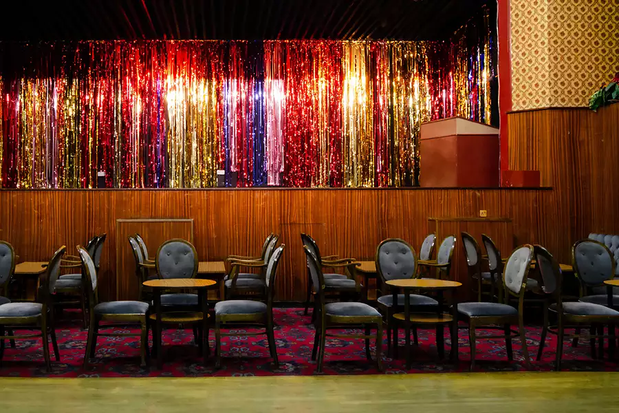 A pub stage with a glittery curtain, blue chairs, tables and a patterned carpeted floor.