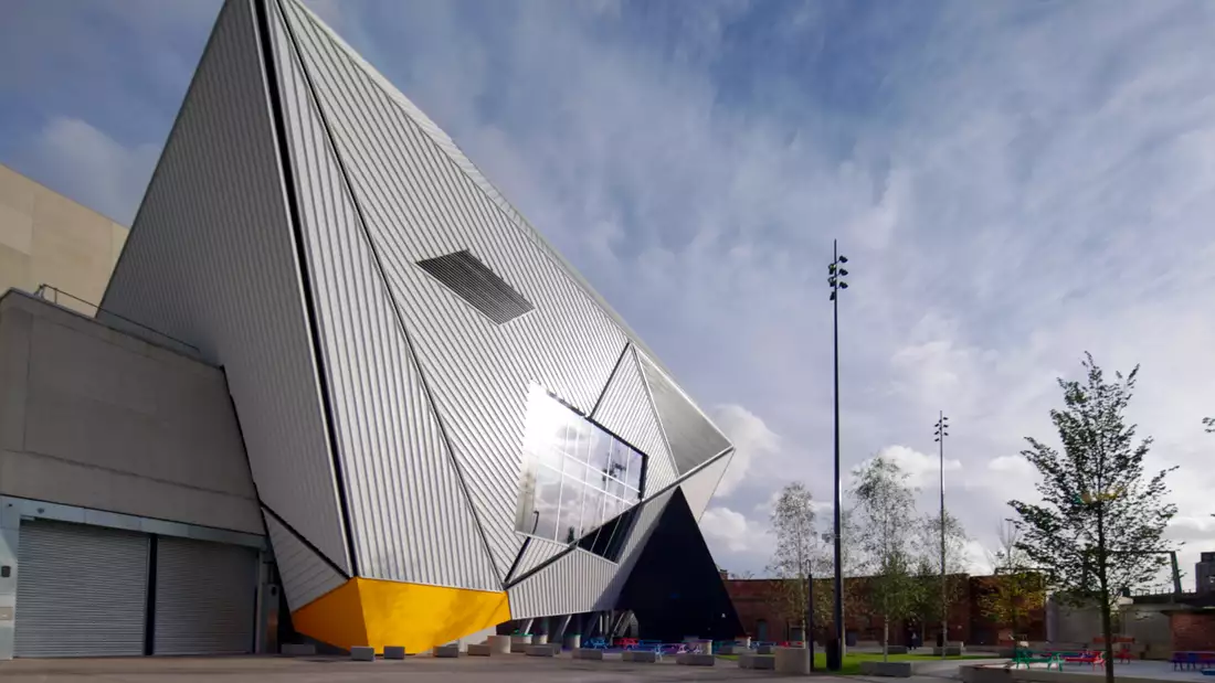 The image shows Aviva Studios, a striking modern building with angular, geometric architecture. The sky above is partly cloudy, and there are some trees and streetlights around the area.