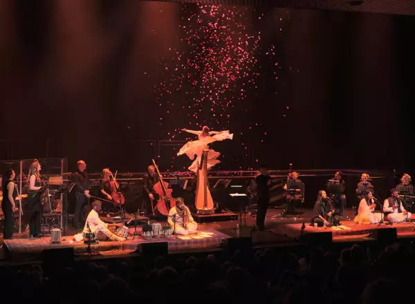A photo of a woman dancing in falling role petals on stage with an orchestra and Sufi singers on the lower level of the stage below her
