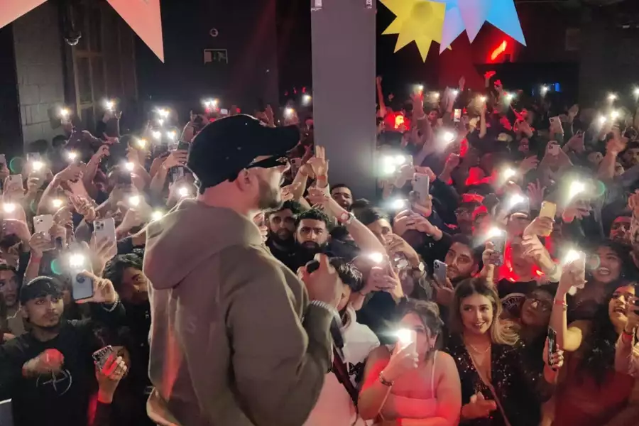 Performer on stage holding a mic, facing a cheering crowd with phone flashlights raised.