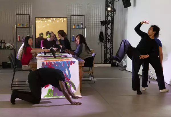 A photo of artist Michael Beard painting on a long roll of paper while dancers from Company Chameleon perform to live music from Manchester Camerata