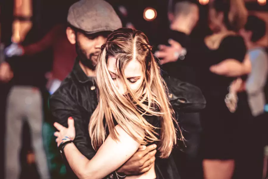 A man and woman are dancing closely in a dimly lit venue, with her long hair partially obscuring her face and his arms wrapped around her. In the blurred background, other couples are also dancing.