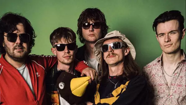 Five members of Madame Claude wearing black shades on a green background. One is holding a large toy penguin.