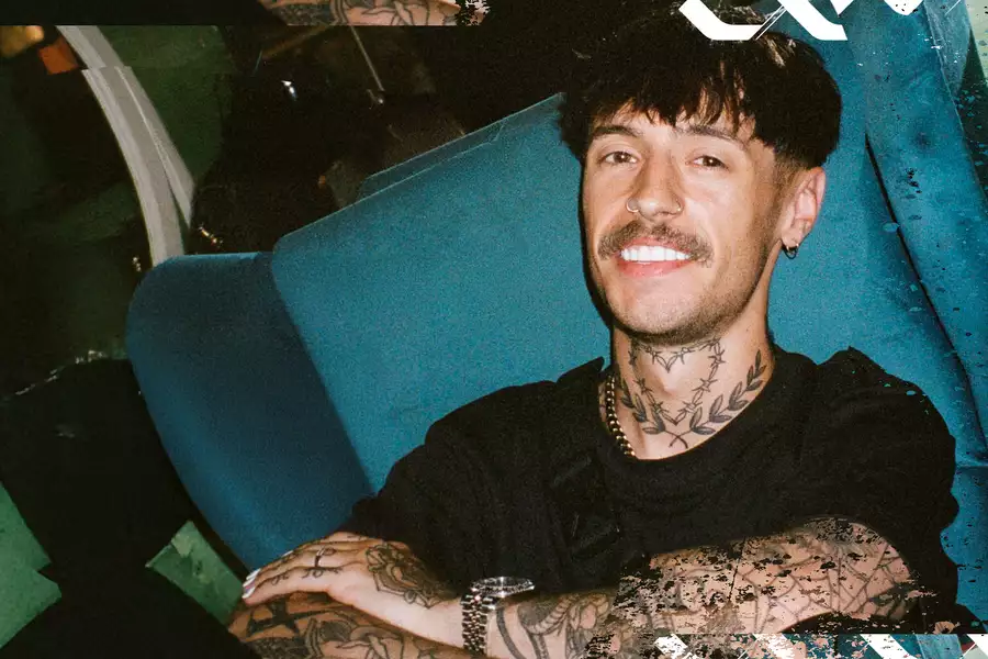 blk. smiling while sitting in a blue chair, wearing a black shirt and silver accessories.