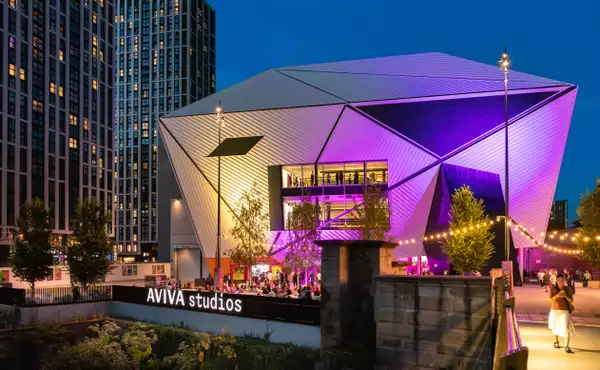 A photo of the venue Aviva Studios lit up with purple lights with crowds enjoying Festival Square by the river
