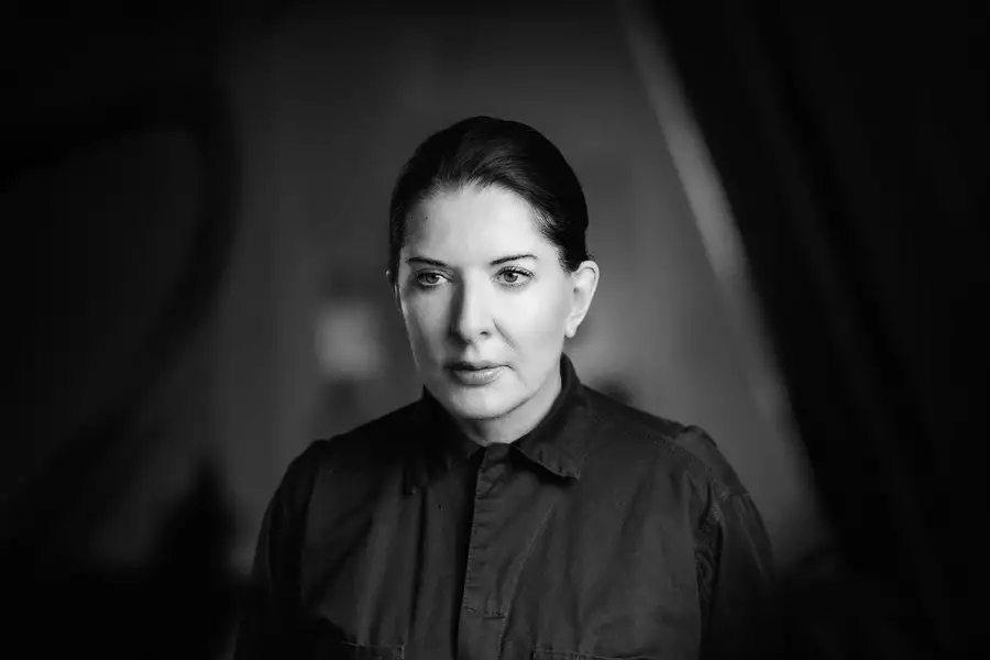 A black-and-white portrait of Marina Abramović, who is shown from the shoulders up, wearing a dark collared shirt. Her hair is neatly pulled back. The background is softly blurred.