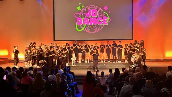 A large group of child dancers on stage underneath a large screen that says 'JD Dance'. They are clapping one performer who is dancing in the middle. There is a crowd of people watching in the audience.