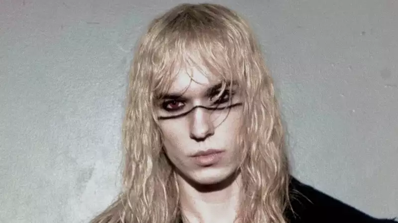 2hollis faces the camera with long blonde hair and bold black eye makeup, wearing a black jacket, striped top and cross necklace against a grey background.