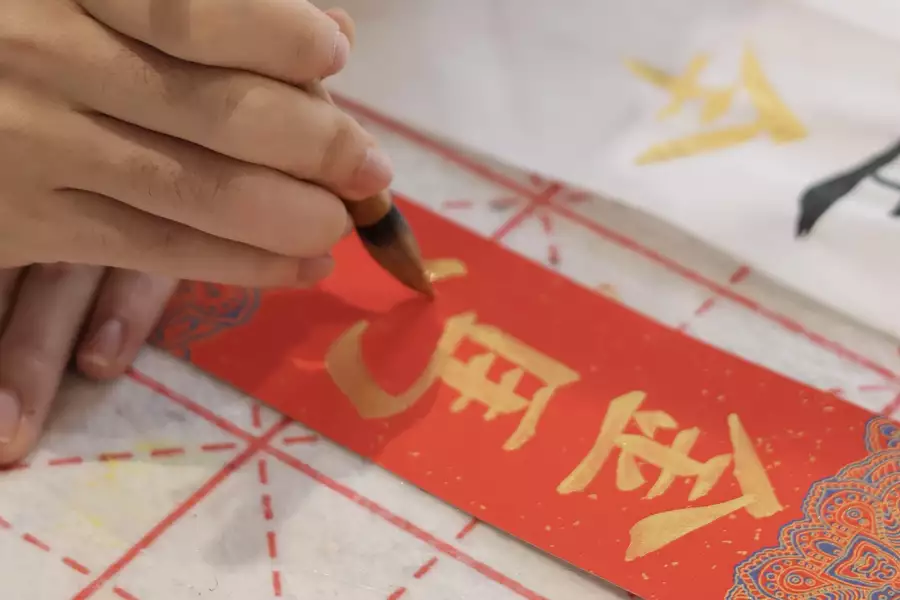 Hand writing Chinese calligraphy in gold ink on a red paper.