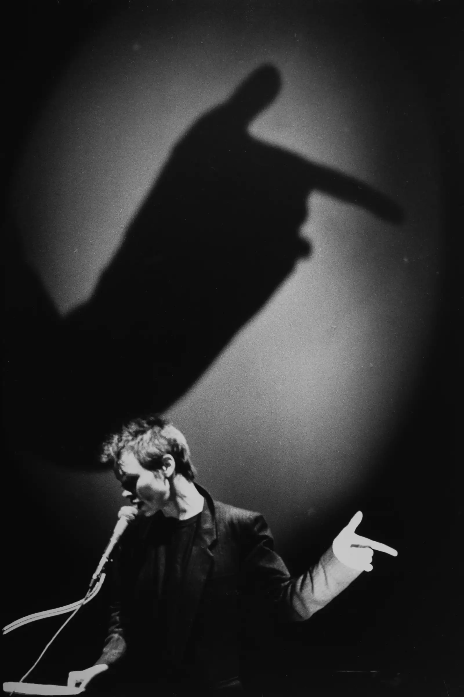 Black and white still from Laurie Anderson's United States Parts 1–4, showing Laurie speaking into a microphone, with a projection of her hand enlarged behind her.