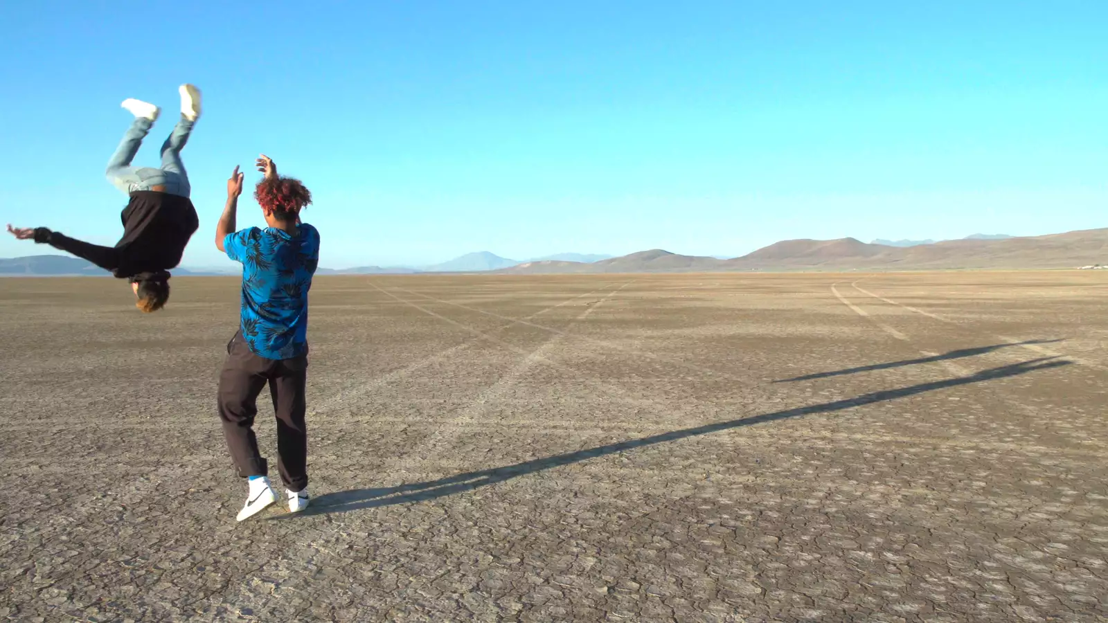 Still from Gasoline Rainbow. Two people on an empty plain – one is doing a backflip, the other is watching with their back turn and clapping. The sky is bright blue and hills are visible in the distance.