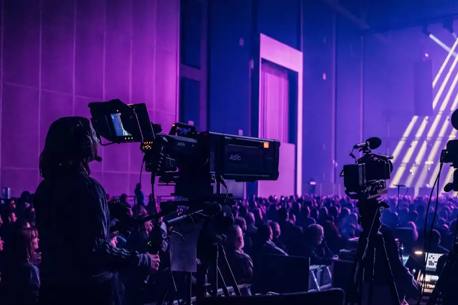 A camera operator films a live event in the Warehouse, which is filled with people. There is dramatic purple lighting with multiple cameras and bright stage beams.