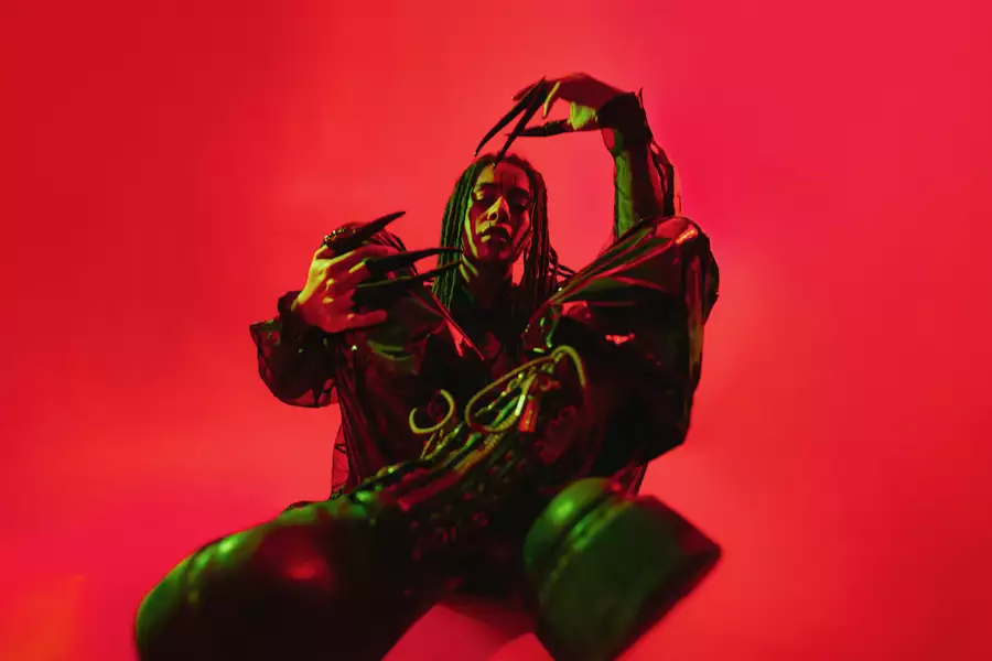 GROVE wearing all black and long black nails, sat crossed legs on a bright red background