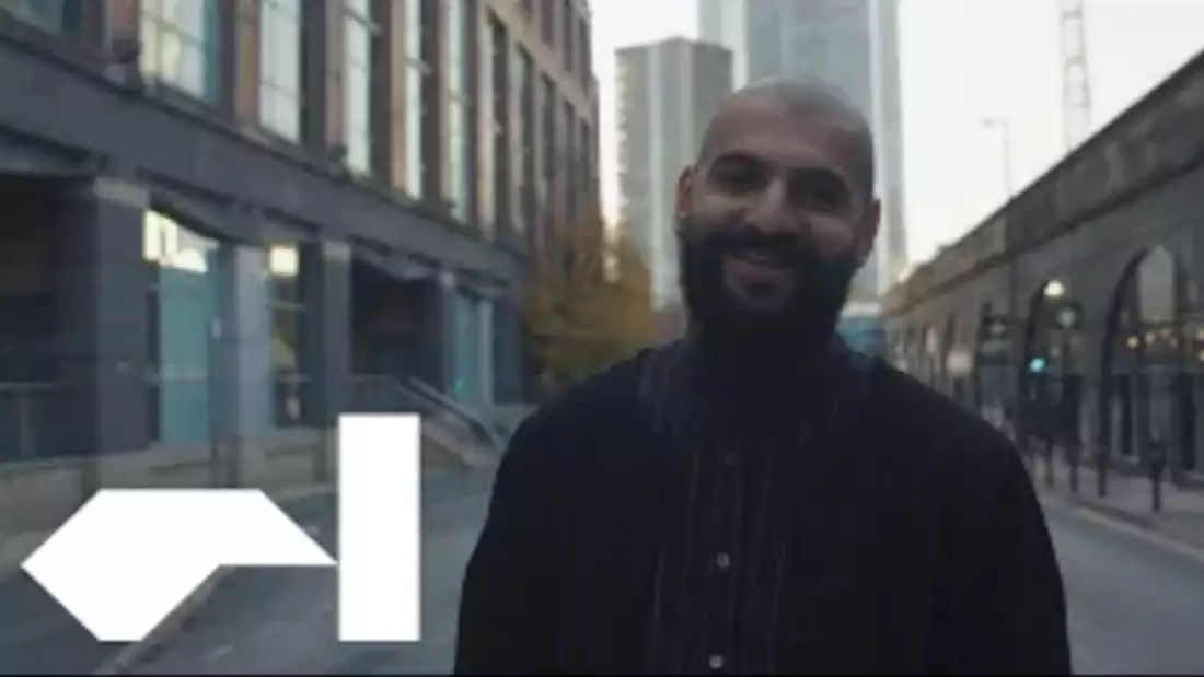 A man with a beard and shaved head smiles towards the camera. He wears a cardigan and striped shirt. In the bottom left hand corner, the Factory International logo is visible.